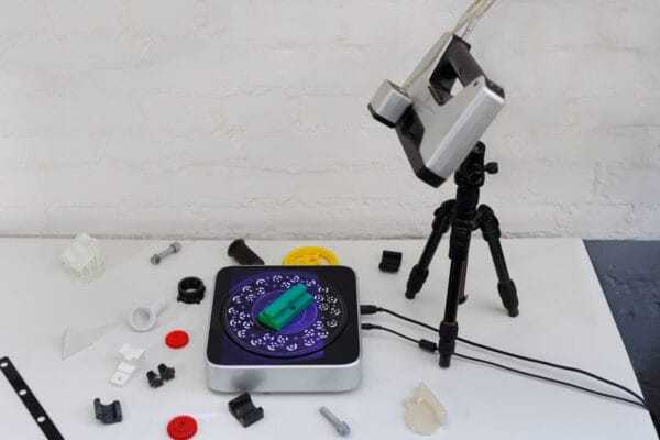 3D Scanner auf einem Stativ vor einem grünen Bauteil auf einem Drehteller mit Referenzmarkierungen.