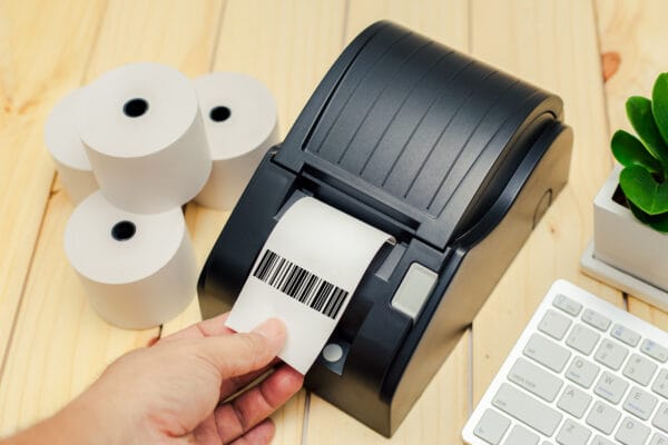 Eine Hand hält ein Barcode-Etikett an der Ausgabe eines schwarzen Etikettendruckers neben weißen Etikettenrollen.