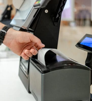 Eine Hand greift nach einem bedruckten Kassenbon, der aus einem schwarzen Belegdrucker an einer Kasse herausragt.