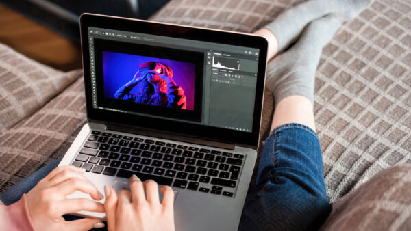 Eine Person sitzt auf einem Sofa und bearbeitet am Laptop ein blau und rot beleuchtetes Porträt einer Person mit VR-Brille.