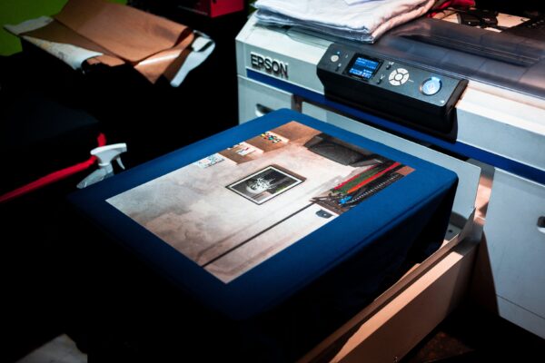 Ein Epson-Textildrucker mit einem blauen T-Shirt und großflächigem Fotomotiv auf der ausgefahrenen Druckplatte.