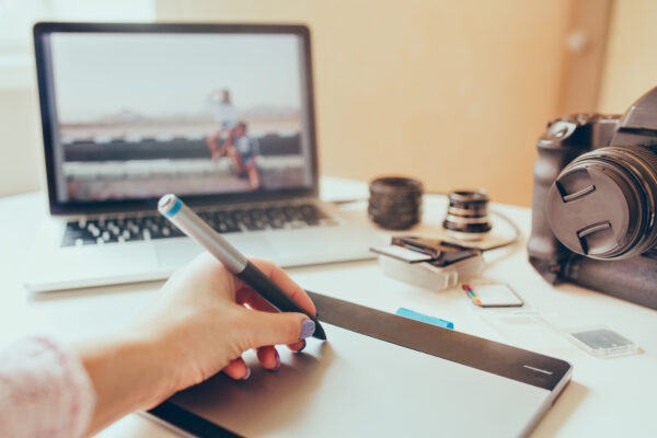 Eine Hand führt einen Stift über ein Grafiktablett; dahinter stehen ein Laptop, eine Kamera, Objektive und Speicherkartenzubehör.