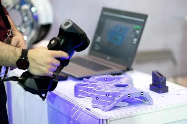 Eine Person führt einen 3D Scanner über ein technisches Bauteil mit Referenzmarkern; dahinter steht ein Laptop.