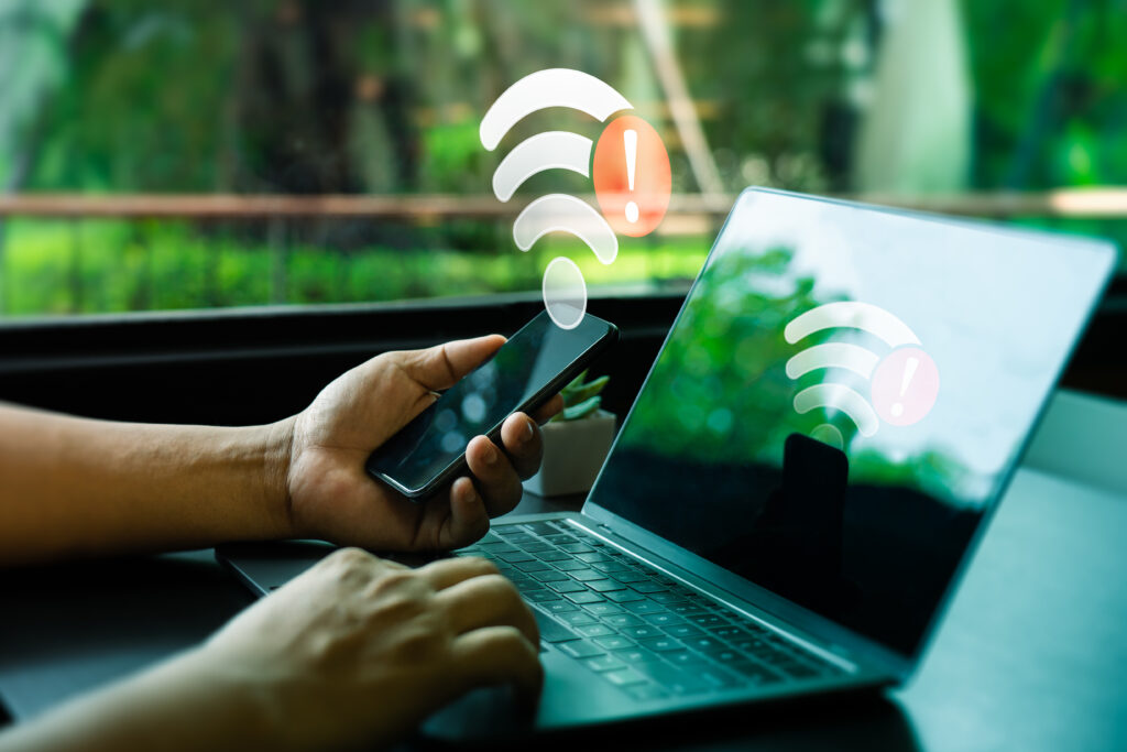 Smartphone und Laptop vor einem Fenster mit eingeblendeten WLAN-Symbolen und Ausrufezeichen als Hinweis auf Verbindungsprobleme.