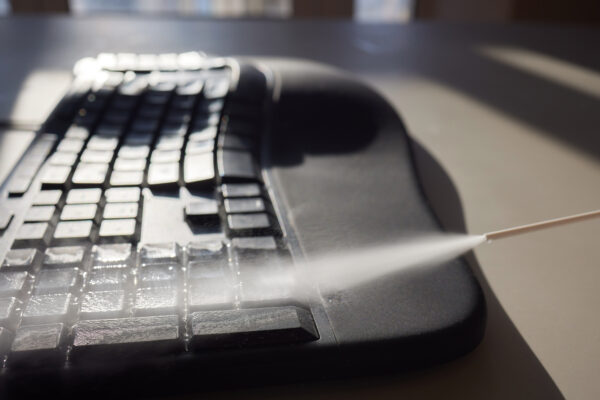 Eine dünne Sprühdüse richtet einen feinen Sprühstrahl auf die Tasten einer schwarzen Computertastatur.